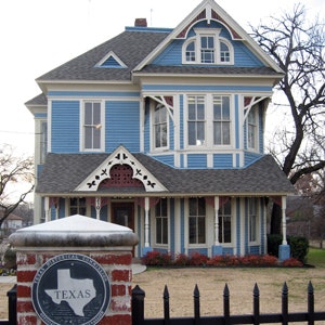 Charming blue Victorian house with elegant white trim and a Texas historical marker welcoming visitors to this picturesque landmark.